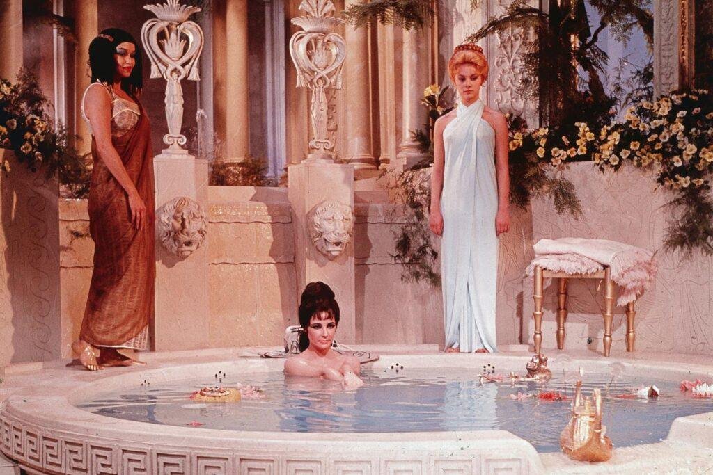 Illustration of Cleopatra VII bathing in milk, adorned with gold jewelry and headdress, representing her ancient beauty rituals.